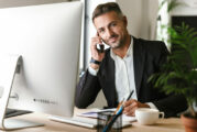 Image of happy businessman talking on cell phone while working on computer in office