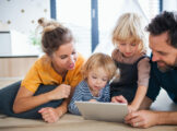 Young family with two small children indoors in bedroom, using tablet.